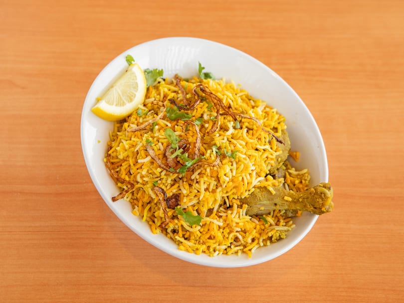 Chicken biryani with yellow rice, fried onions, cilantro, and a lemon wedge in a white bowl.